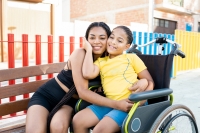 A black girl with braided hair has fun with her brother sitting in a wheelchair in a park.The young people hug each other while looking at the camera happily.