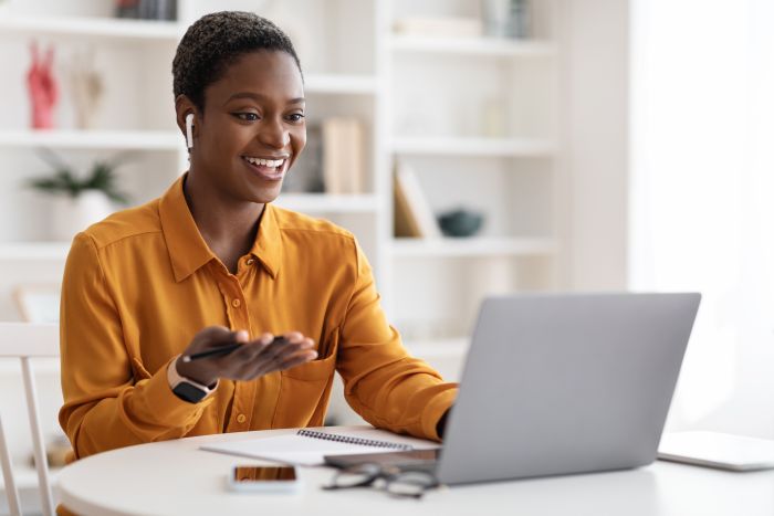Attractive young African American woman having video conference with clients or business partners, sitting at workdesk at office, using laptop and earbuds, gesturing and smiling,