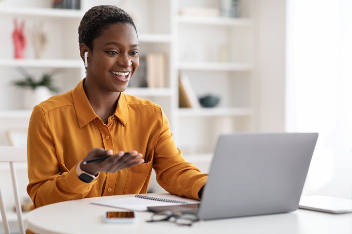 Attractive young African American woman having video conference with clients or business partners, sitting at workdesk at office, using laptop and earbuds, gesturing and smiling,